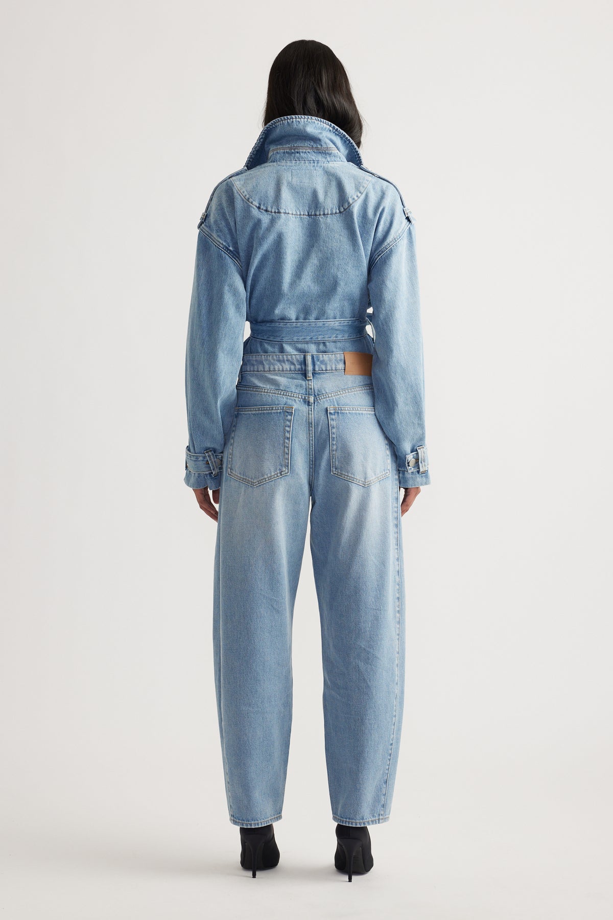 Woman shown from behind wearing light indigo cropped denim jacket with high-waisted barrel leg jeans and black heels