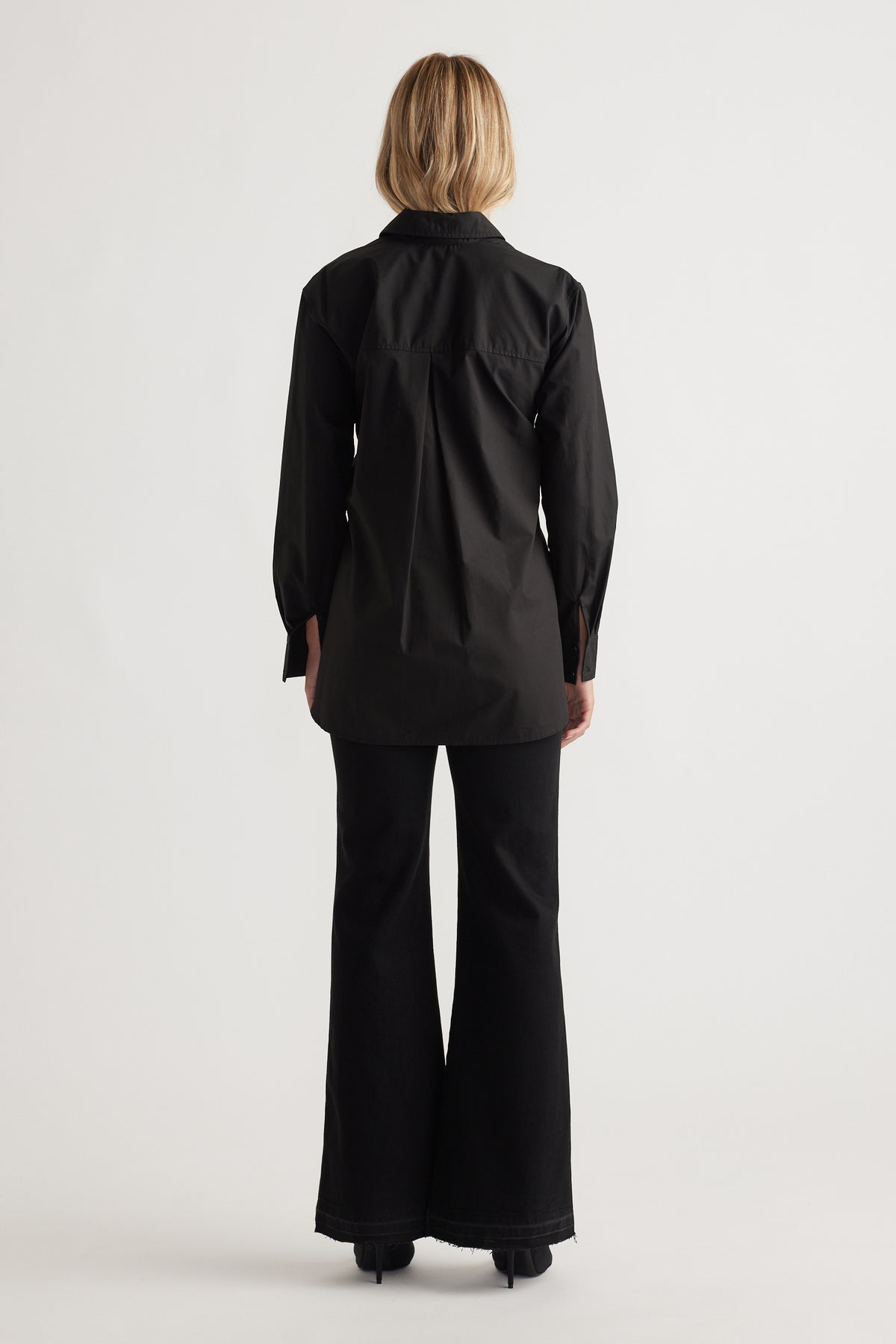 Back view of a woman wearing a sleek black tailored shirt with long sleeves paired with flared black pants and heels