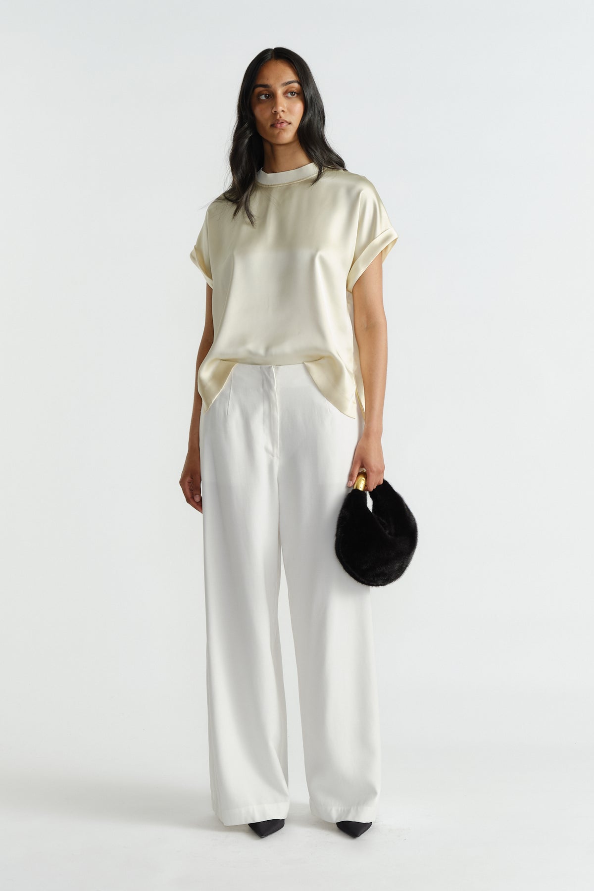 Woman wearing a relaxed satin tee in buttery cream paired with wide-leg white pants and holding a black faux fur bag.