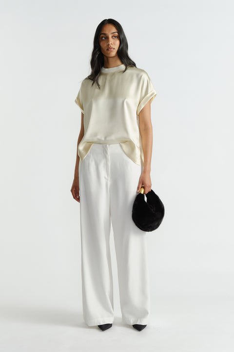 Woman wearing a relaxed satin tee in buttery cream paired with wide-leg white pants and holding a black faux fur bag.