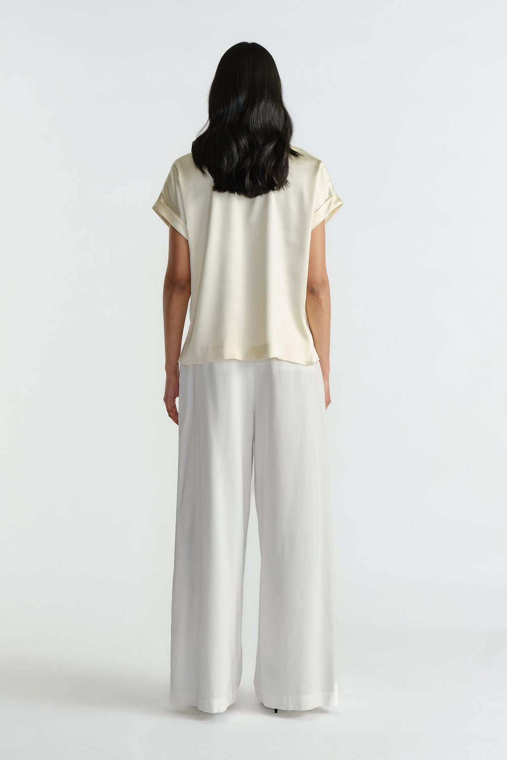 Woman wearing a relaxed fit satin short sleeve top in creamy beige paired with flowing wide-leg white pants.