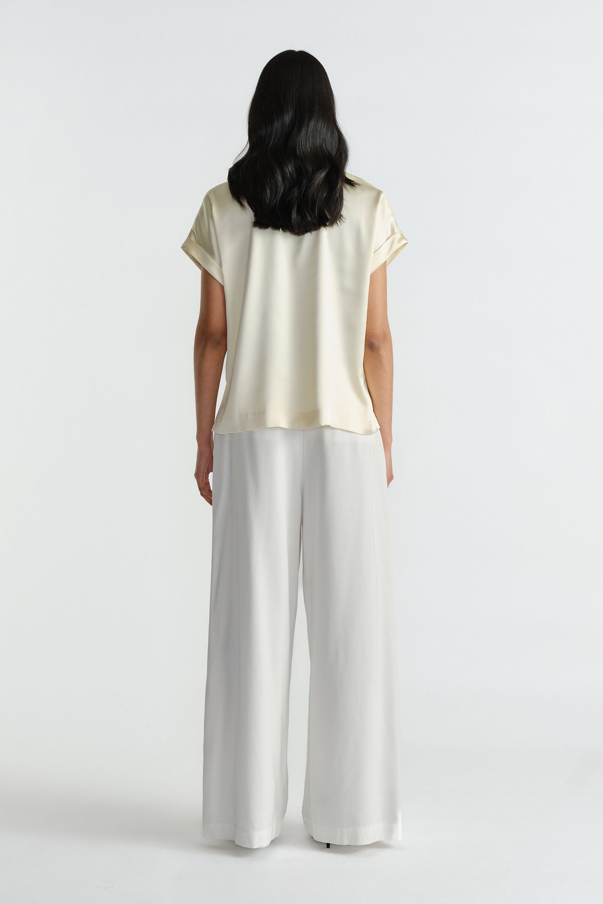 Woman wearing a relaxed fit satin short sleeve top in creamy beige paired with flowing wide-leg white pants.