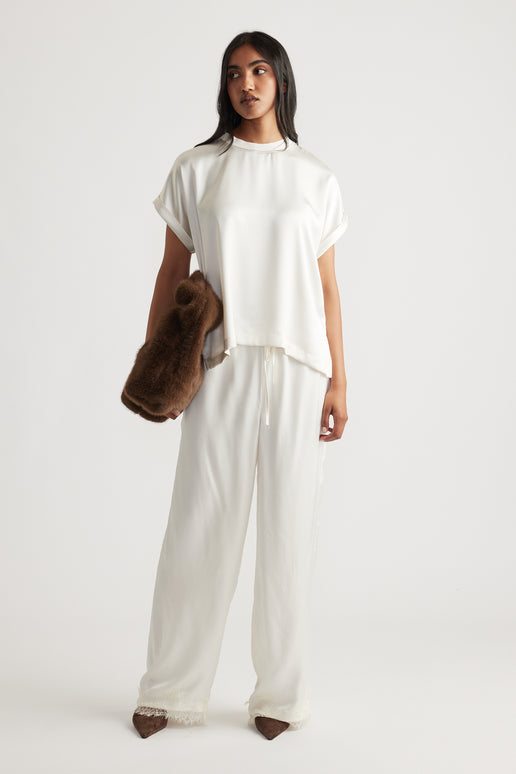 Woman wearing an ivory satin relaxed tee paired with matching loose trousers and holding a soft brown furry clutch
