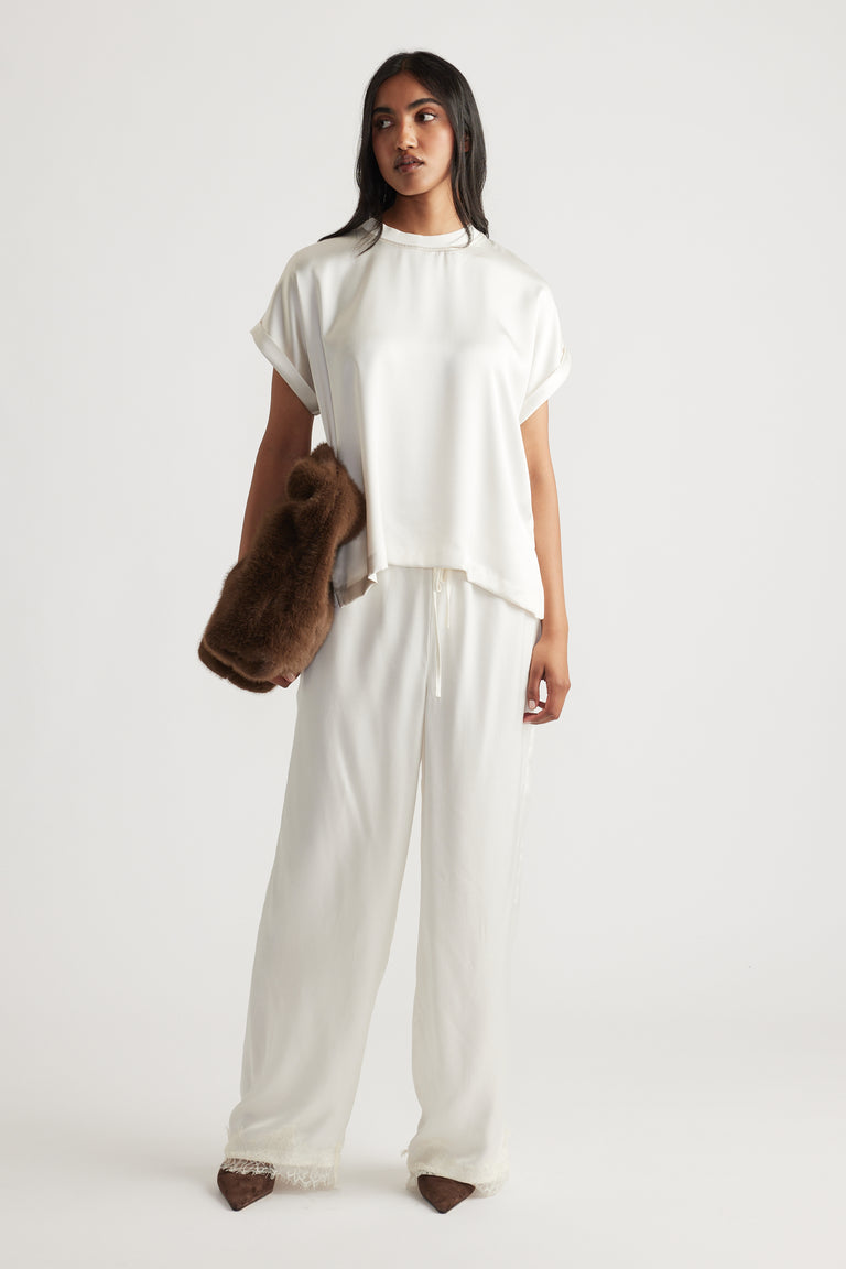 Woman wearing an ivory satin relaxed tee paired with matching loose trousers and holding a soft brown furry clutch