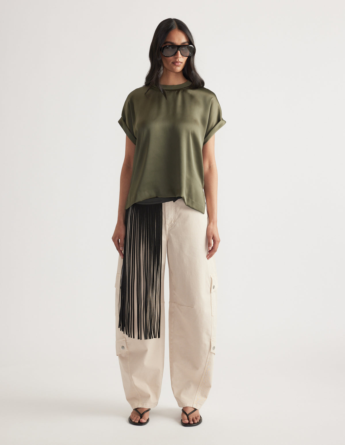 Woman wearing an olive satin short sleeve top paired with cream wide-leg pants featuring black fringe detail and sandals