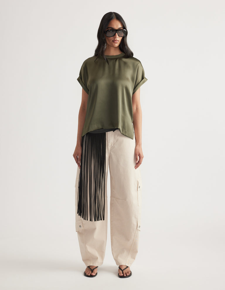 Woman wearing an olive satin short sleeve top paired with cream wide-leg pants featuring black fringe detail and sandals