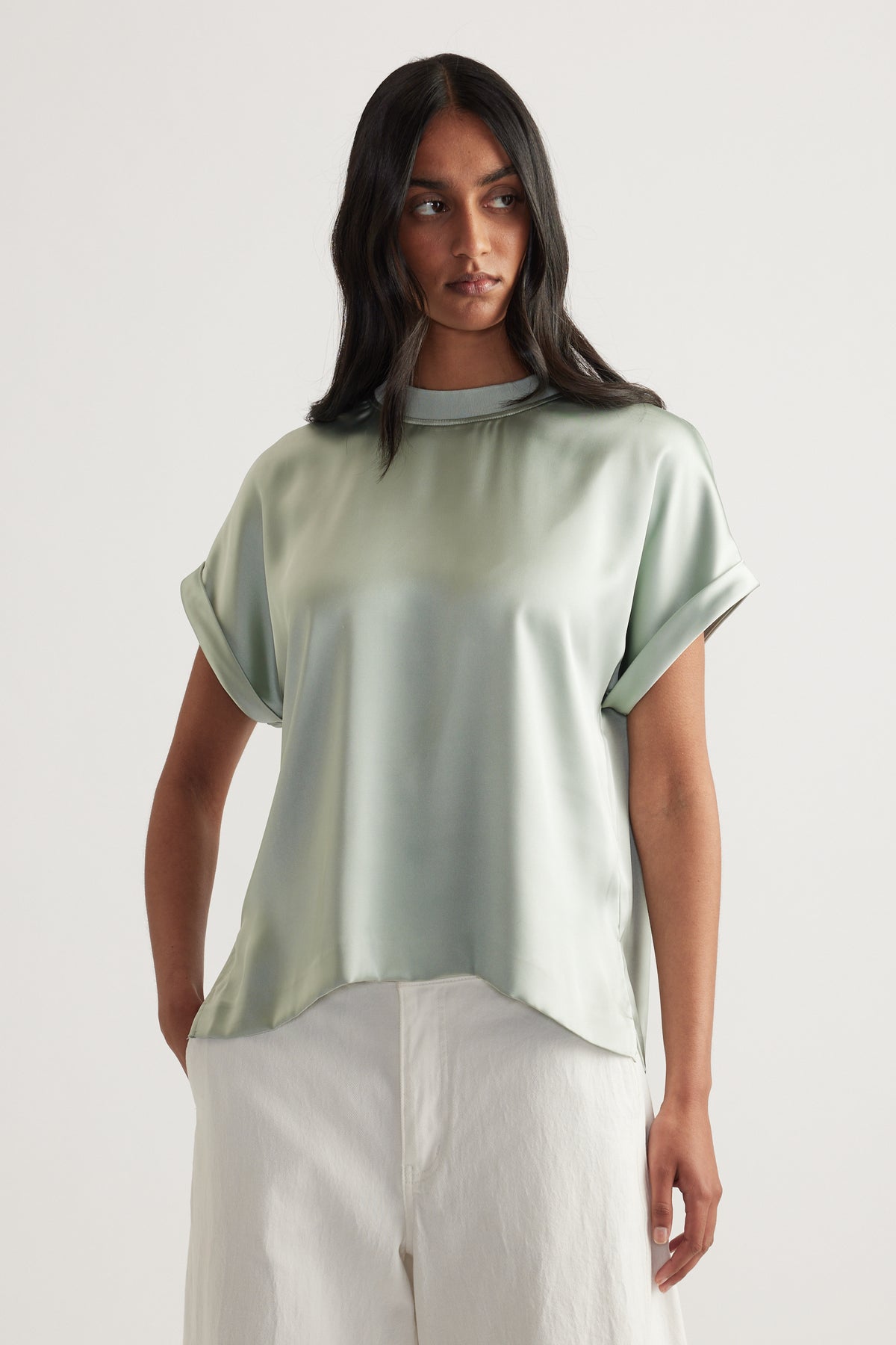 Woman wearing a sage green satin crew neck tee with rolled sleeves and white pants