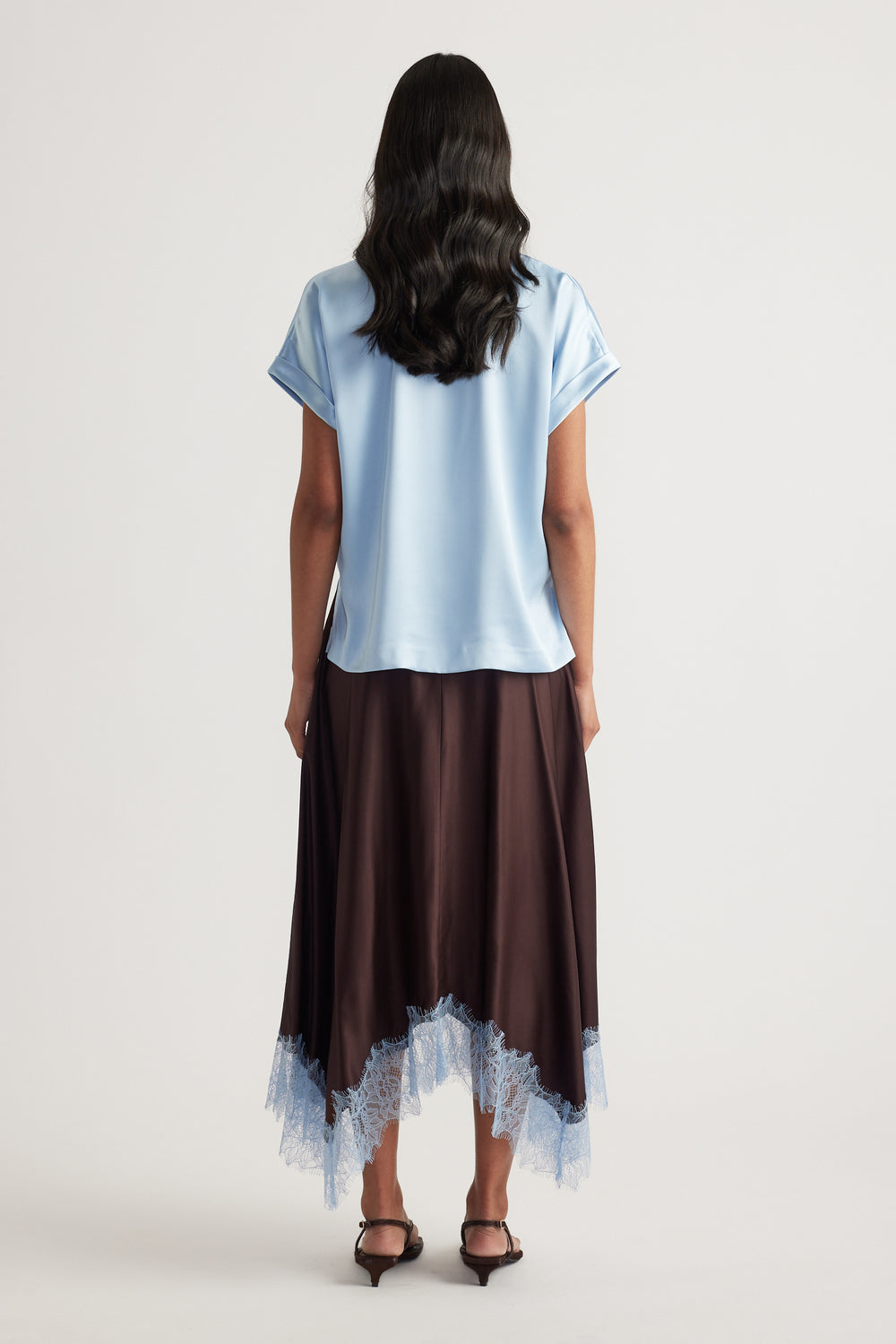 Woman viewed from behind wearing a light blue satin tee with a dark lace hem skirt and black heels
