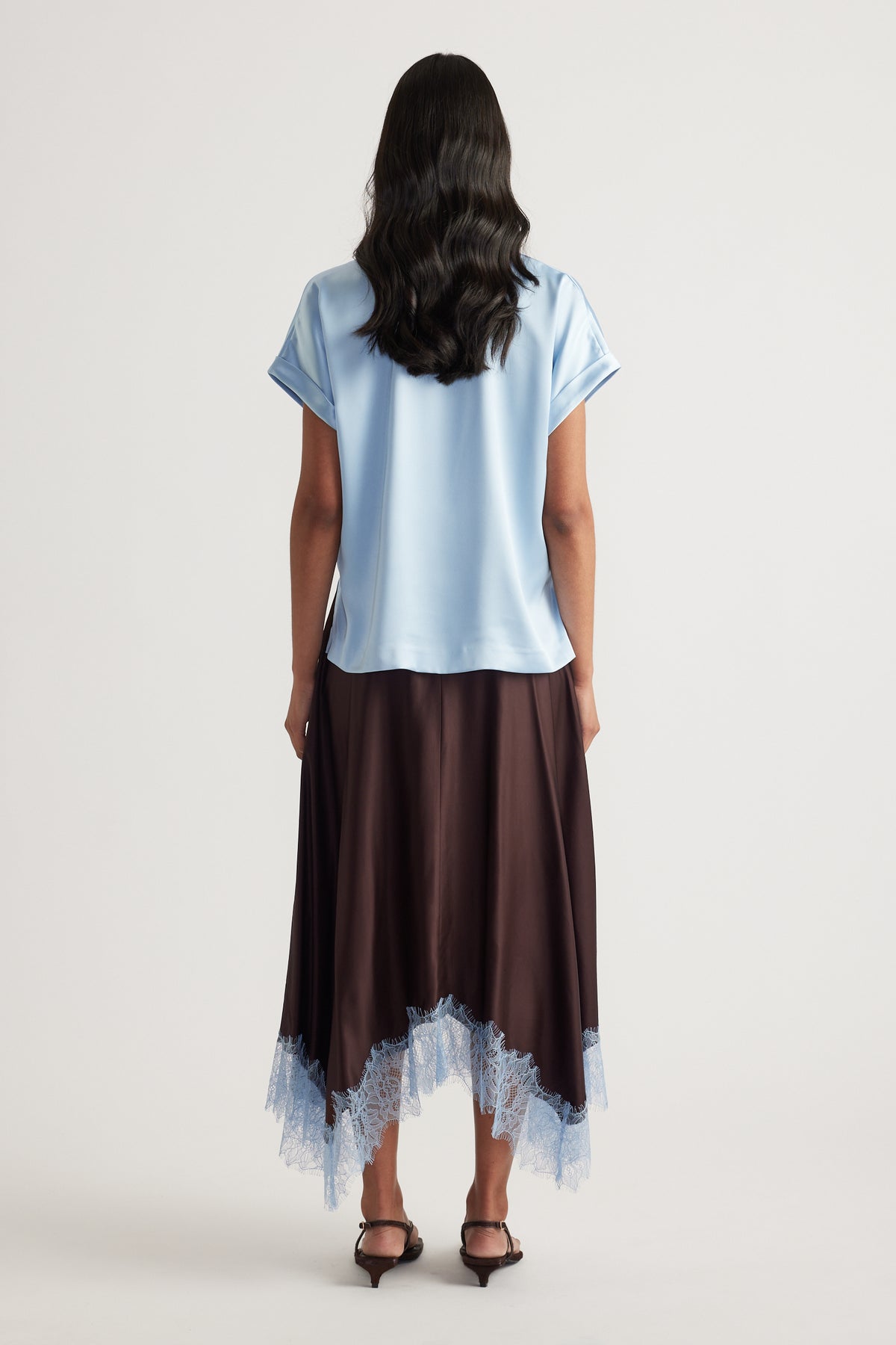 Woman viewed from behind wearing a light blue satin tee with a dark lace hem skirt and black heels