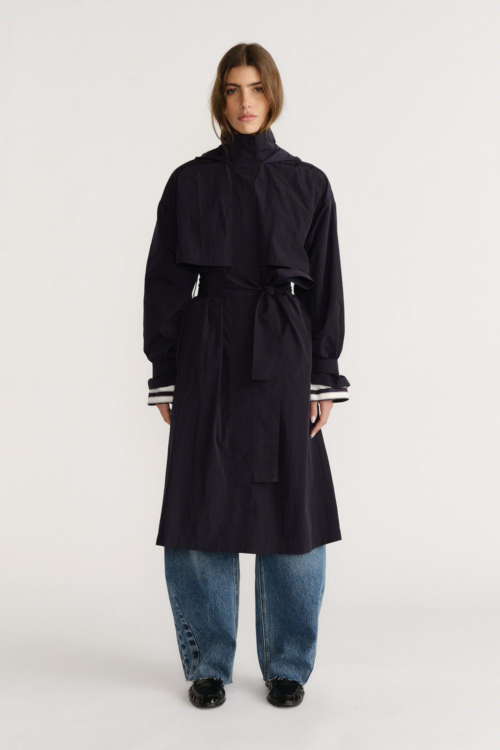 Woman wearing a navy belted trench coat with high collar over loose blue jeans and black shoes, posed front view.