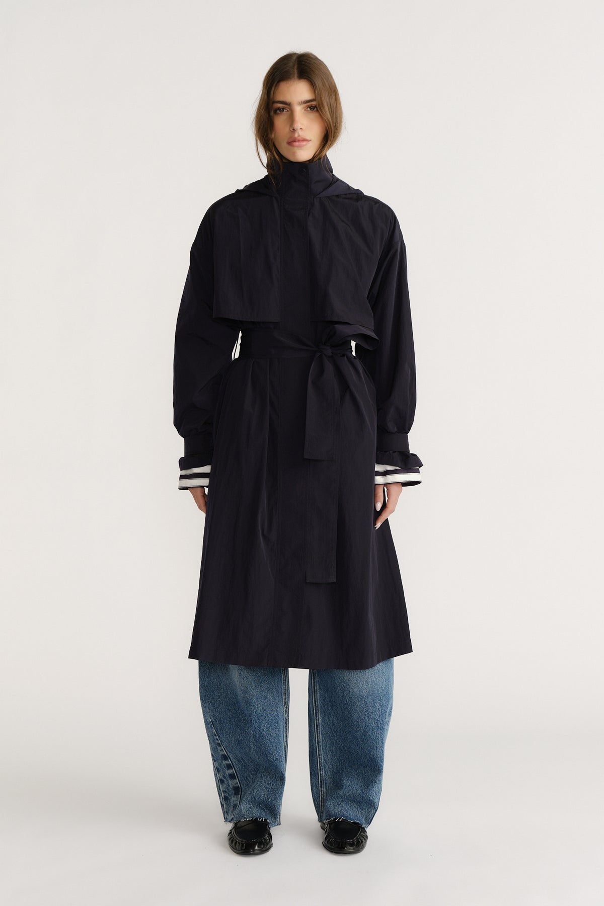 Woman wearing a navy belted trench coat with high collar over loose blue jeans and black shoes, posed front view.