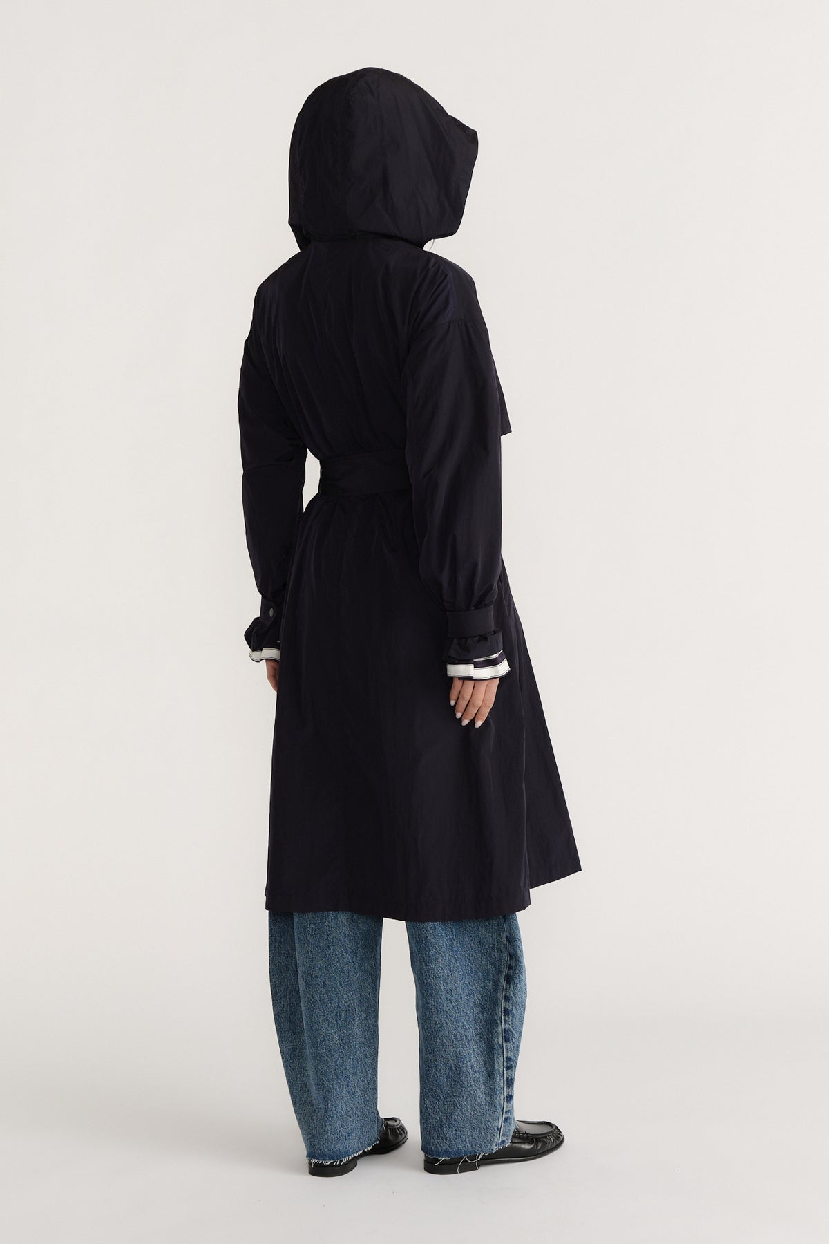 Woman wearing a navy lightweight trench coat with hood and belt, paired with relaxed blue jeans and black shoes viewed from behind