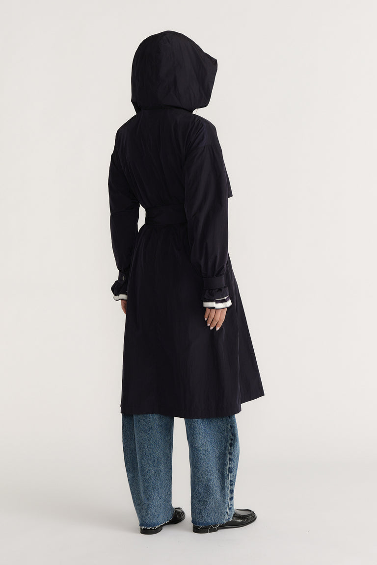 Woman wearing a navy lightweight trench coat with hood and belt, paired with relaxed blue jeans and black shoes viewed from behind