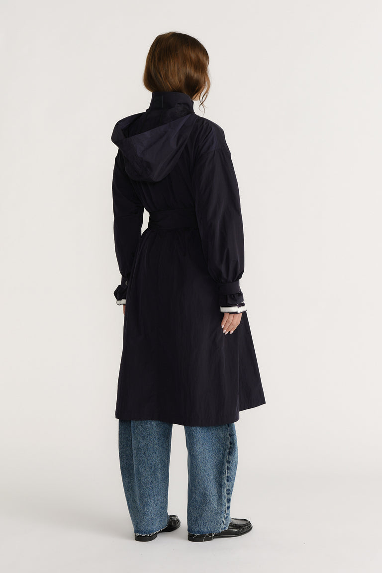 Woman wearing a navy long trench coat with a hood and belt, paired with loose blue jeans and black shoes, seen from behind.