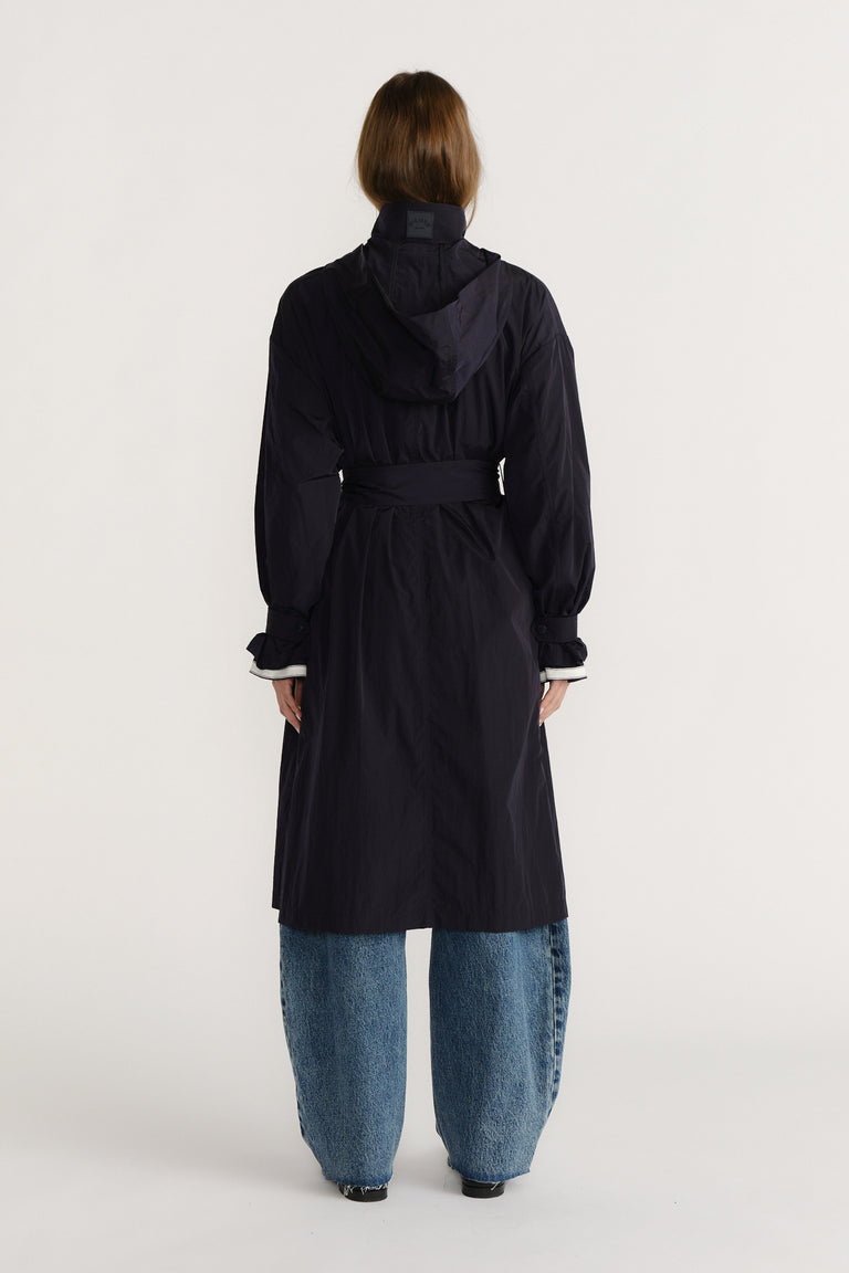 Rear view of woman wearing a navy belted trench coat with a hood paired with wide-leg blue jeans and black shoes