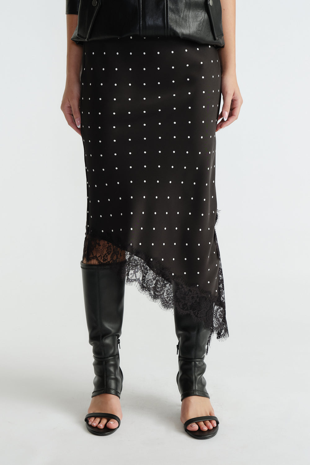 Front view of black midi slip skirt with white polka dots, asymmetric lace trim hem, worn with black boots and jacket