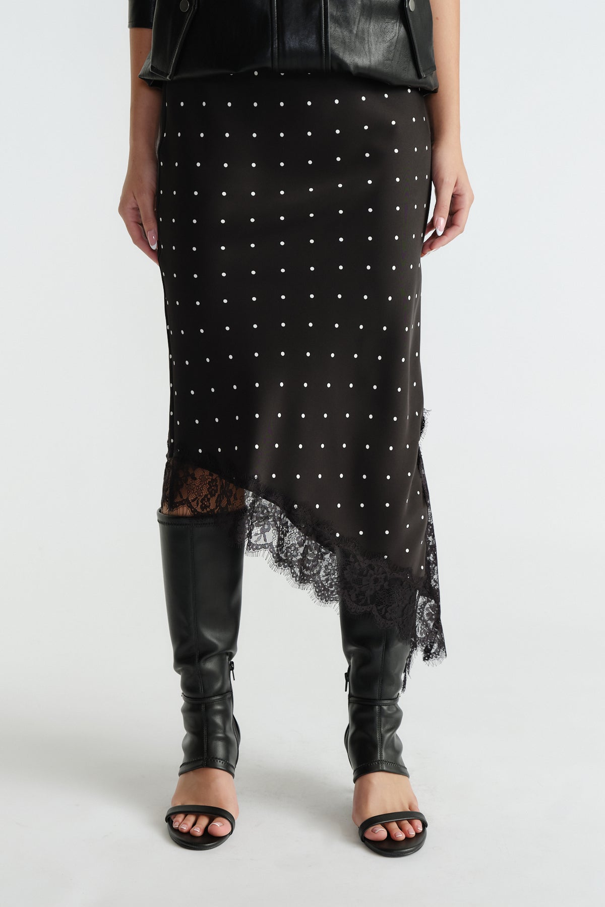 Front view of black midi slip skirt with white polka dots, asymmetric lace trim hem, worn with black boots and jacket
