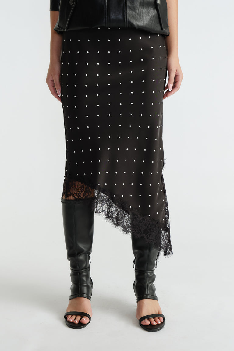 Front view of black midi slip skirt with white polka dots, asymmetric lace trim hem, worn with black boots and jacket