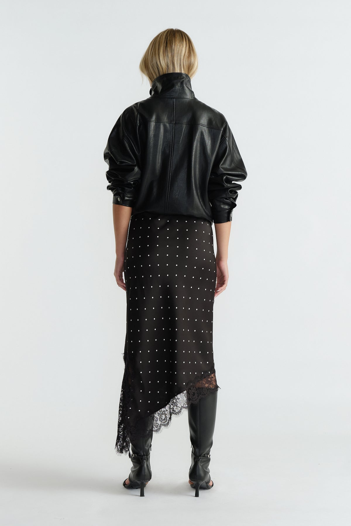 Rear view of woman wearing black asymmetric hem midi skirt with lace trim and studded detail paired with black jacket