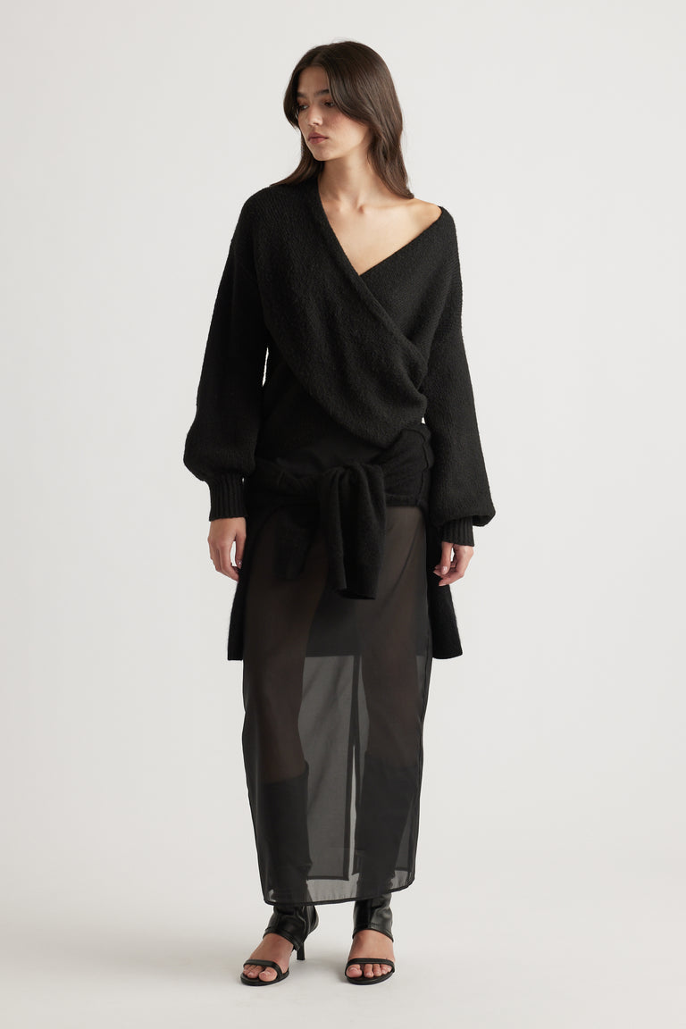Woman wearing a textured black crossover jumper with balloon sleeves over sheer black maxi skirt and open-toe heels.