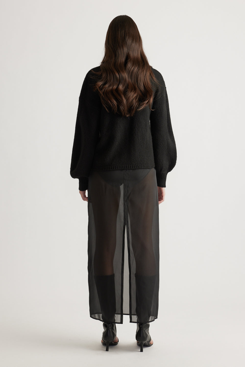 Rear view of a woman wearing a black jumper with balloon sleeves and sheer black wide-leg pants with ankle boots.