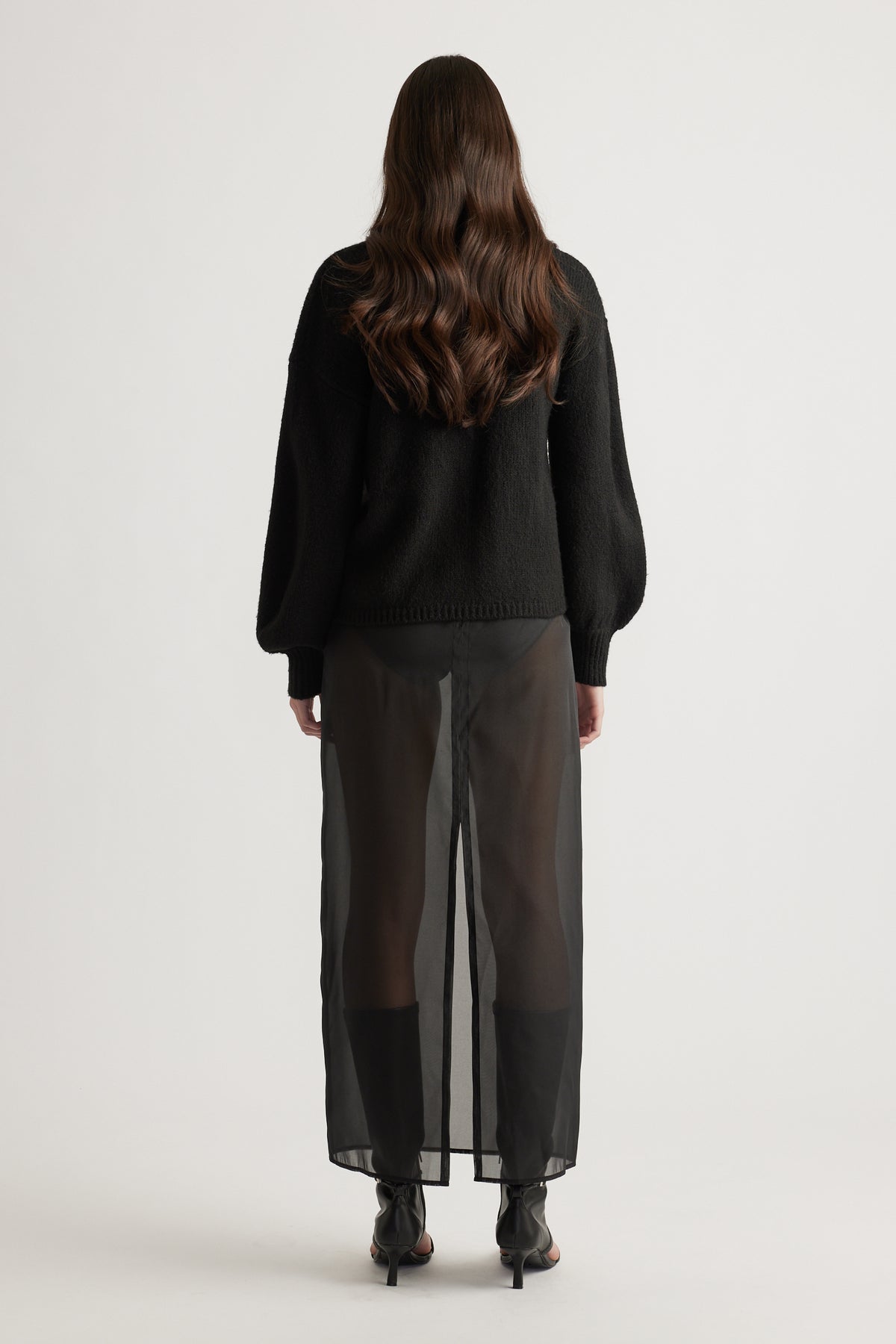 Rear view of a woman wearing a black jumper with balloon sleeves and sheer black wide-leg pants with ankle boots.