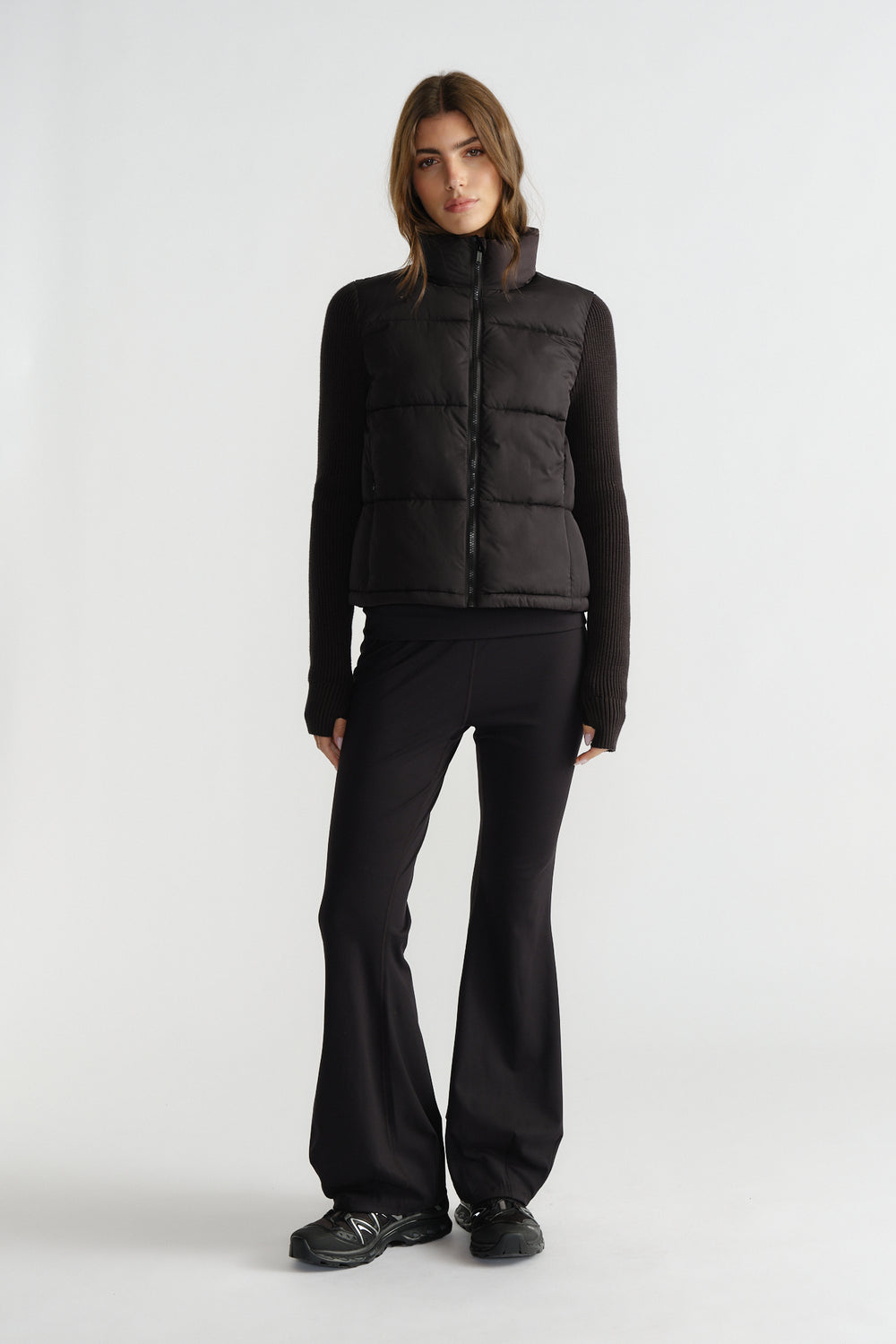 Woman wearing a black knit puffer jacket layered over a fitted long sleeve top with flared black pants and sneakers.