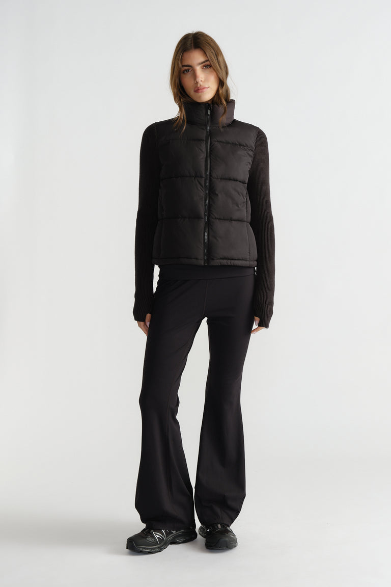 Woman wearing a black knit puffer jacket layered over a fitted long sleeve top with flared black pants and sneakers.