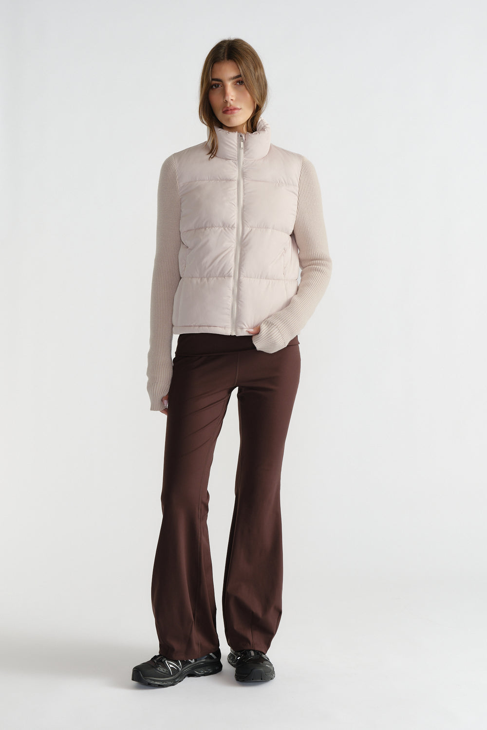 Woman wearing a light beige knit puffer jacket with ribbed sleeves paired with dark brown flared pants and black sneakers