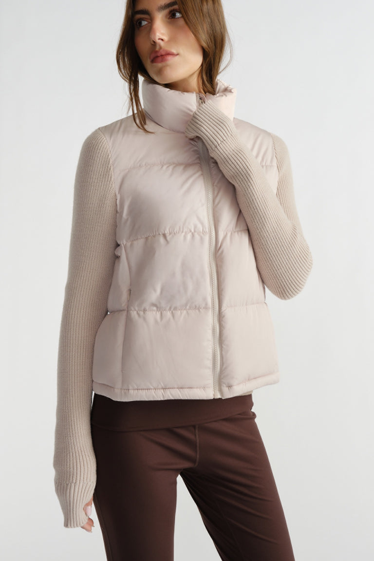 Woman wearing a light beige knit-sleeve puffer vest with high collar paired with brown fitted pants