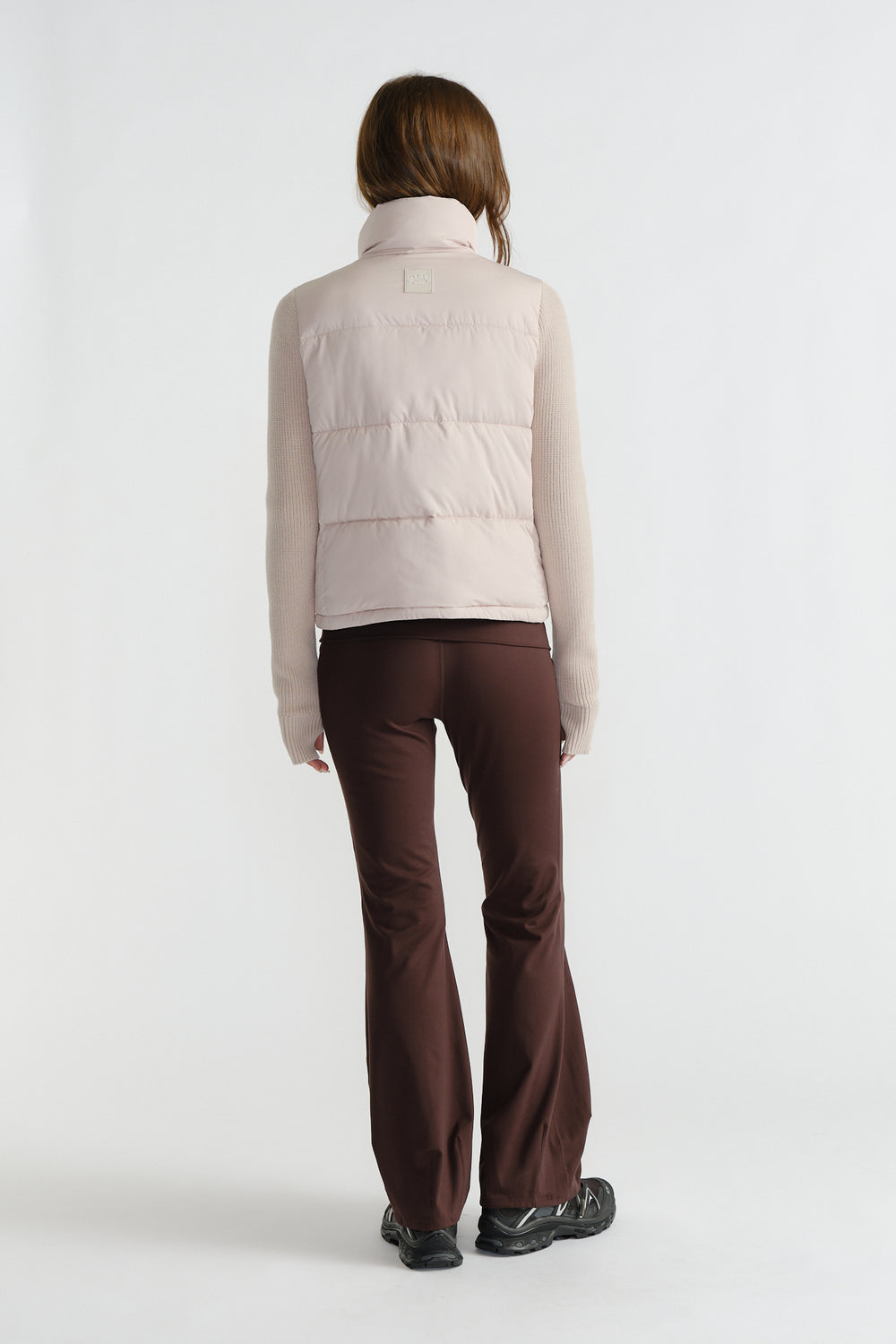 Back view of woman wearing a light pink knit puffer jacket with ribbed sleeves and dark brown flared pants paired with black shoes