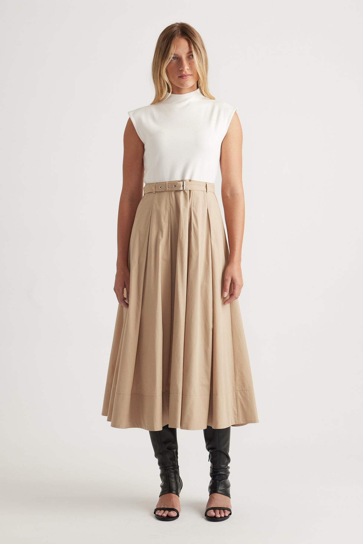 Woman wearing Stefanie Spliced Midi Dress in warm tan with belted waist, structured pleats, styled with black boots and heels