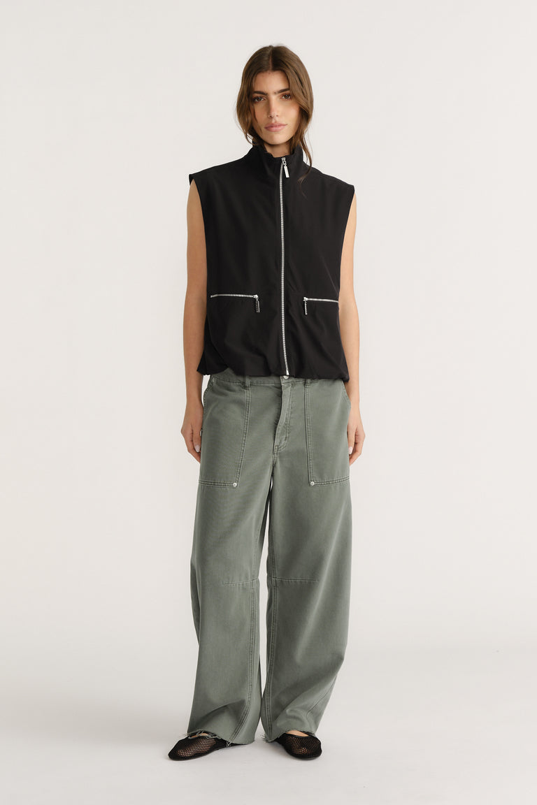 Woman wearing a sleeveless black zip-up vest with high collar paired with loose wide-leg olive green pants and black flats