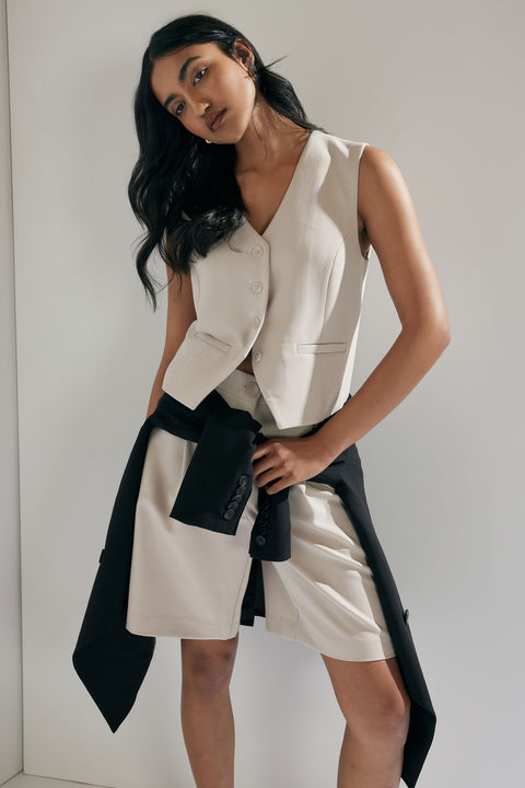 Woman in tailored cream vest and matching shorts with black jacket tied around her waist in a minimal setting