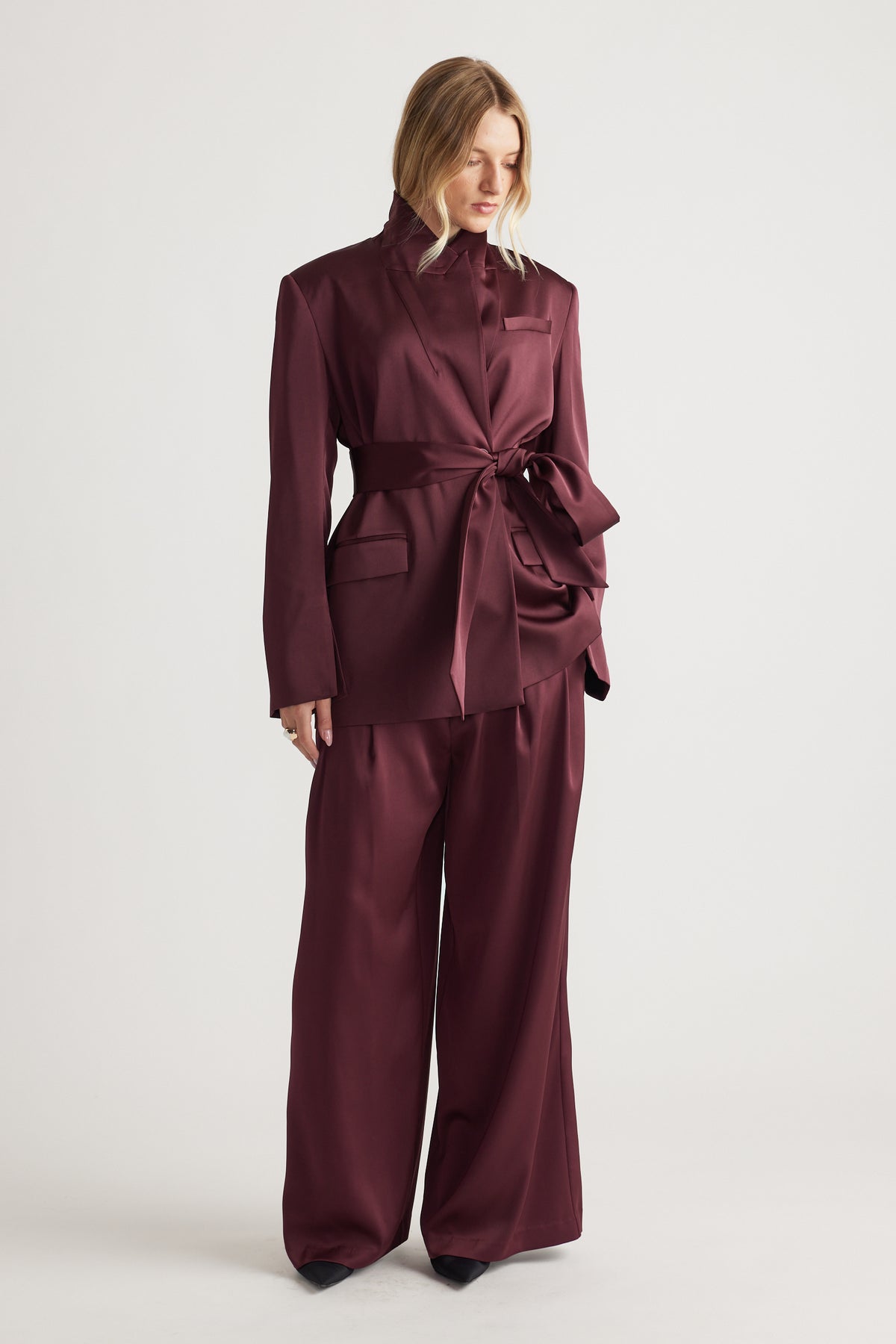 Woman wearing a deep plum satin wrap blazer with a tied waist paired with wide-leg matching trousers