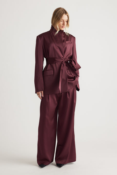 Woman wearing a deep plum satin wrap blazer with a tied waist paired with wide-leg matching trousers