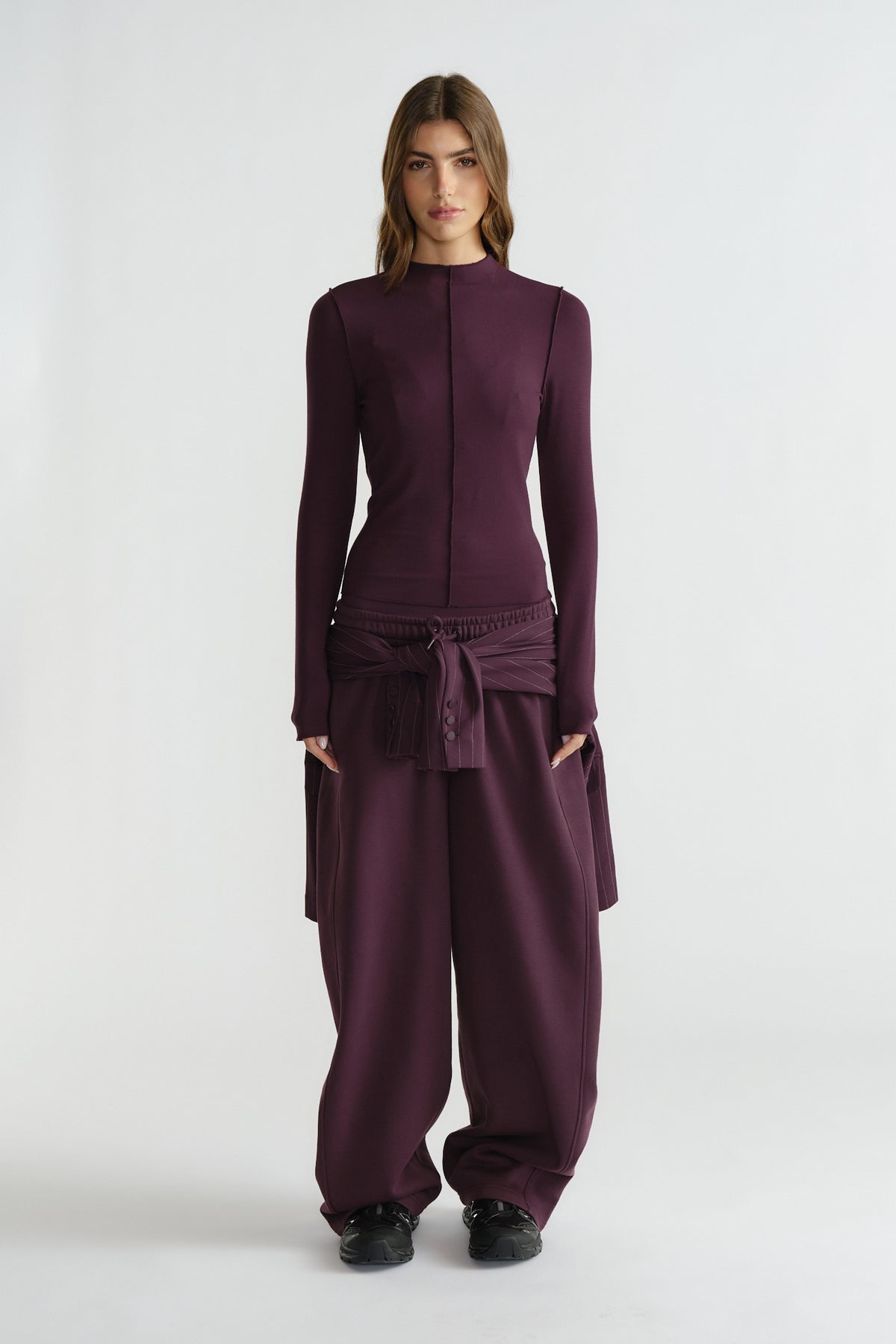 Woman wearing a fitted long sleeve top and loose wide-leg pants in deep plum with a tied waist detail and black shoes