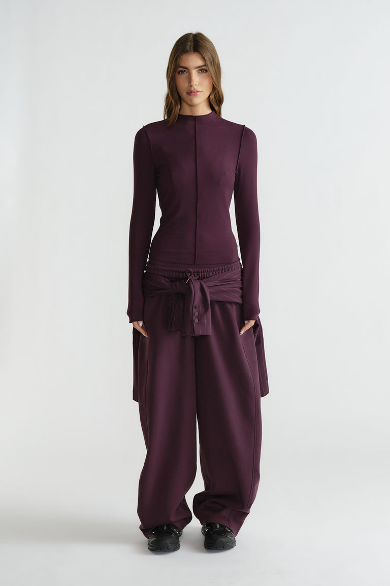 Woman wearing a fitted long sleeve top and loose wide-leg pants in deep plum with a tied waist detail and black shoes