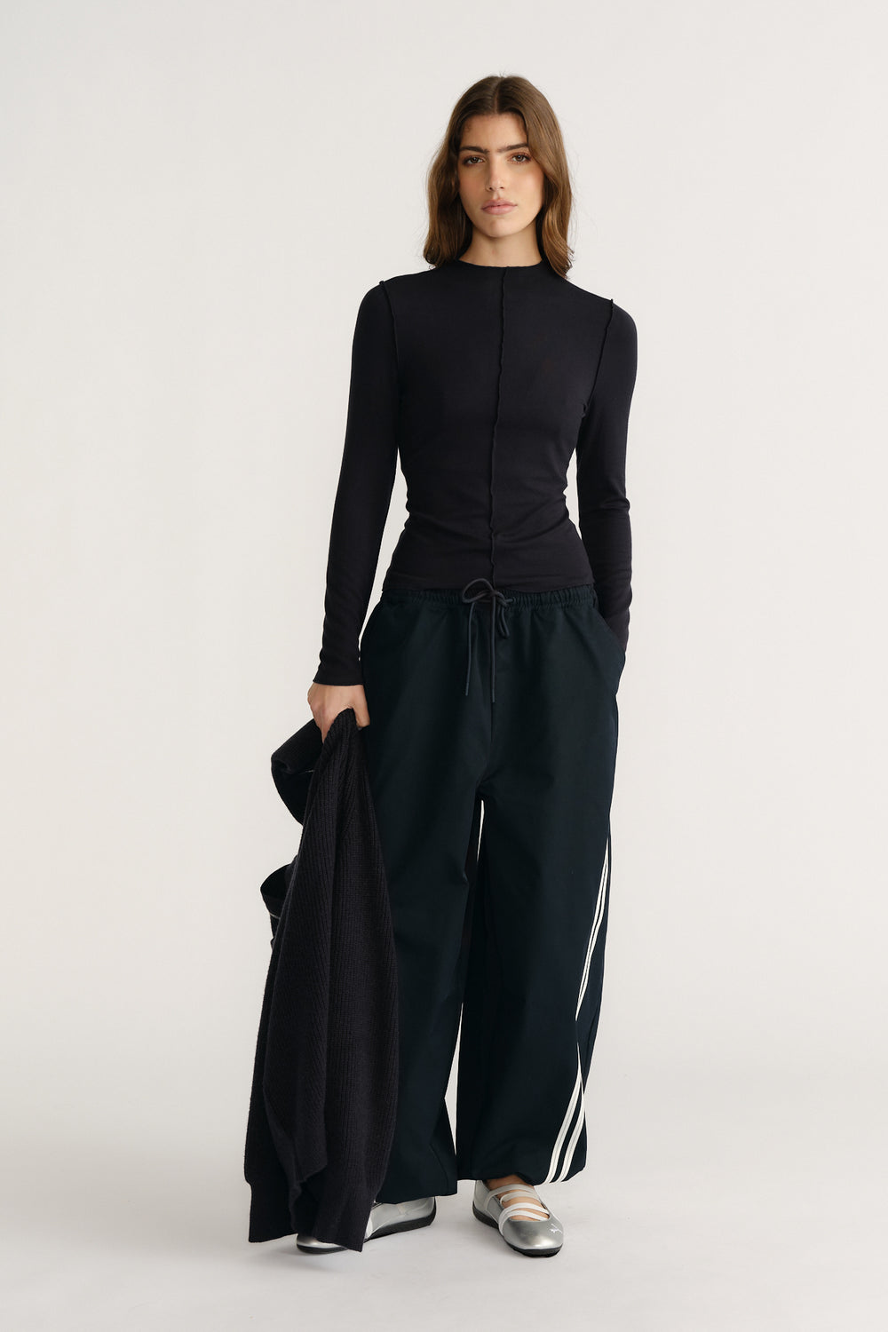 Woman wearing navy long sleeve fitted top paired with wide-leg track pants and metallic silver flats, holding a cardigan.