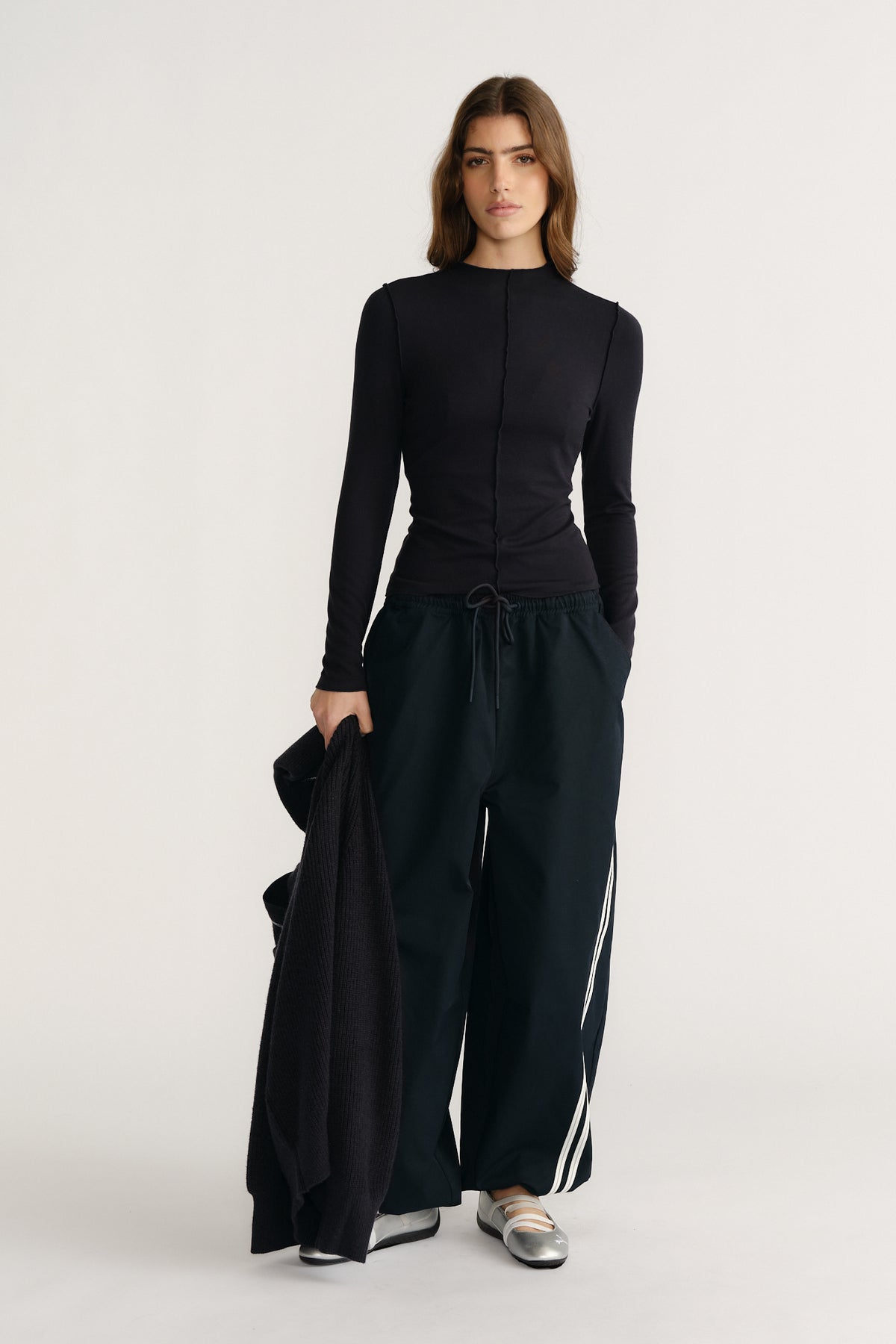 Woman wearing navy long sleeve fitted top paired with wide-leg track pants and metallic silver flats, holding a cardigan.