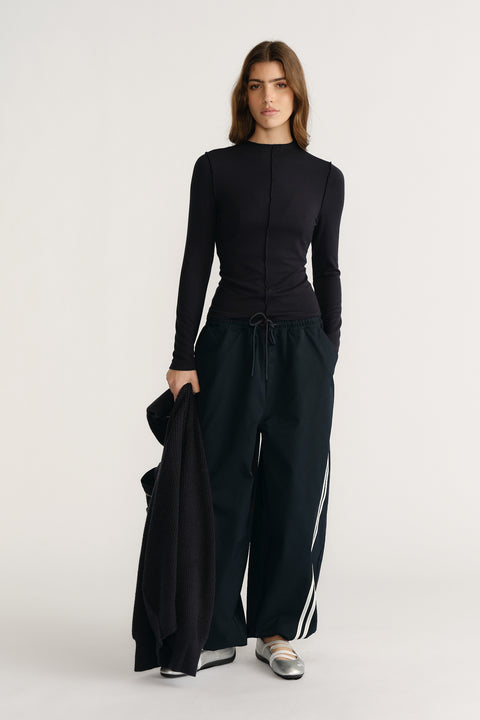 Woman wearing navy long sleeve fitted top paired with wide-leg track pants and metallic silver flats, holding a cardigan.