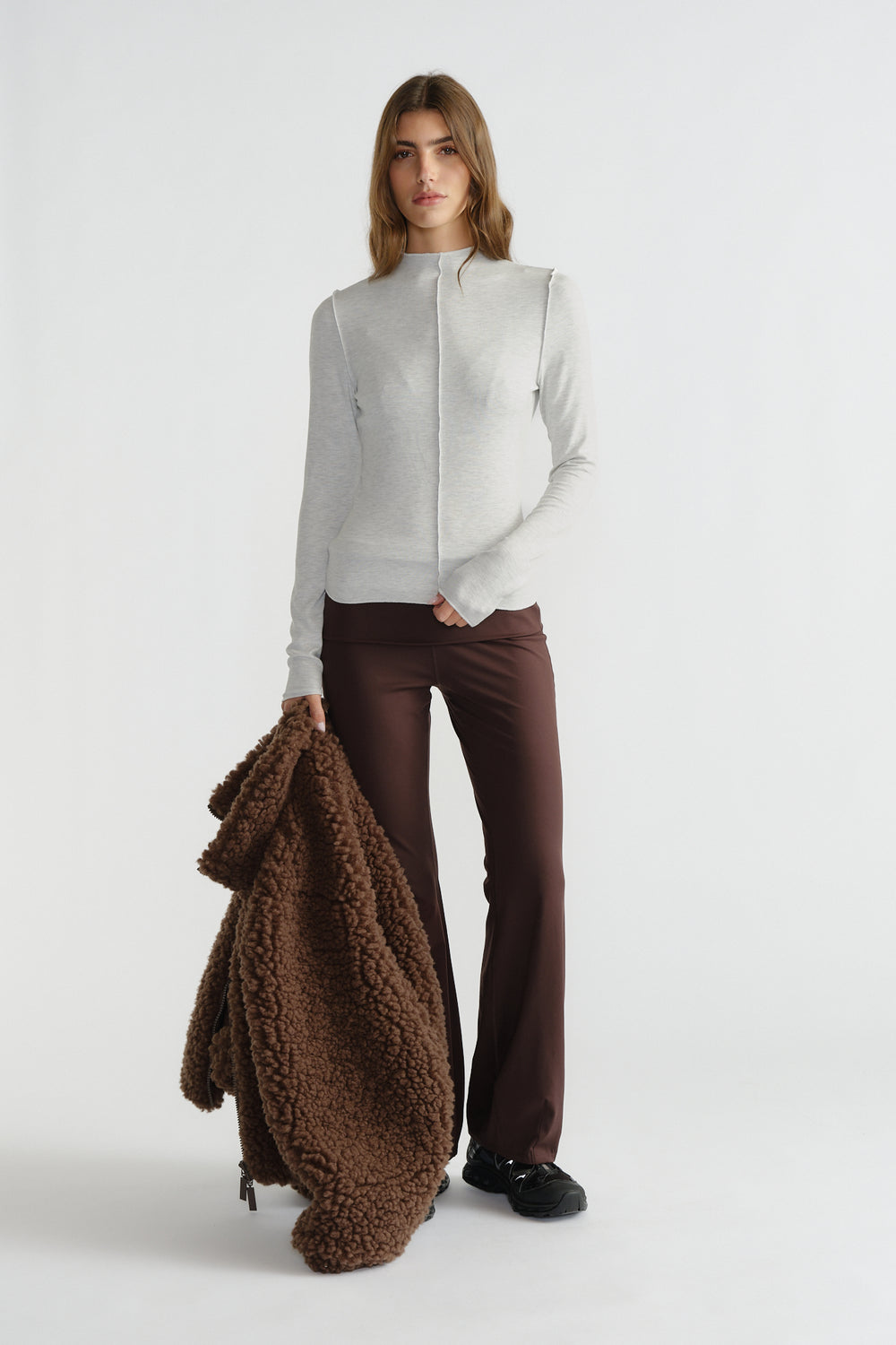 Woman in light grey fitted long sleeve top and chocolate brown flared pants holding a textured brown jacket