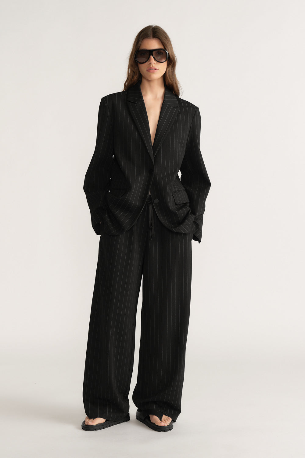 Woman wearing black pinstripe wide-leg pants with matching oversized blazer and dark sunglasses on plain background
