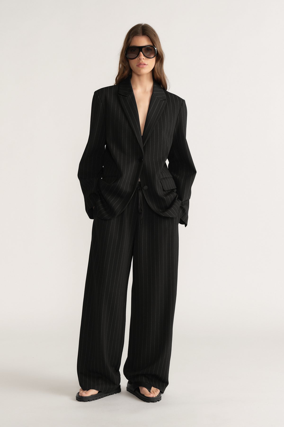 Woman wearing black pinstripe wide-leg pants with matching oversized blazer and dark sunglasses on plain background