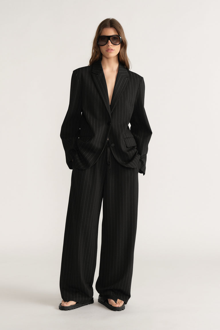 Woman wearing black pinstripe wide-leg pants with matching oversized blazer and dark sunglasses on plain background