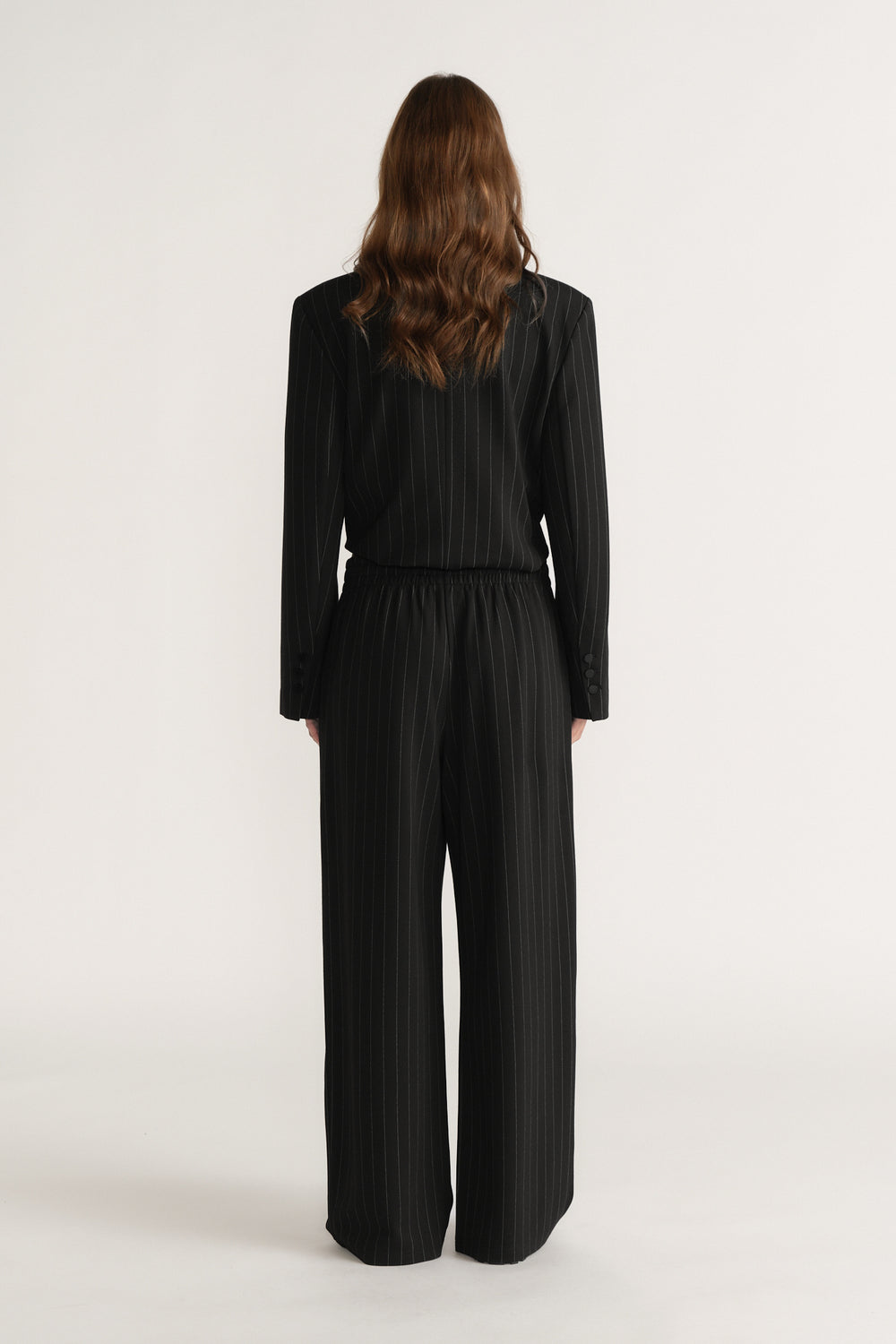 Woman wearing black pinstripe wide-leg pants and matching long-sleeve top, viewed from the back on plain background