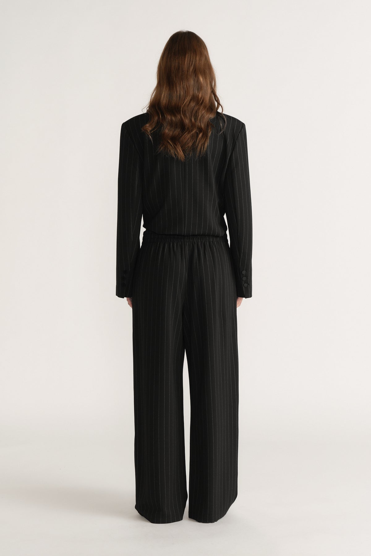 Woman wearing black pinstripe wide-leg pants and matching long-sleeve top, viewed from the back on plain background
