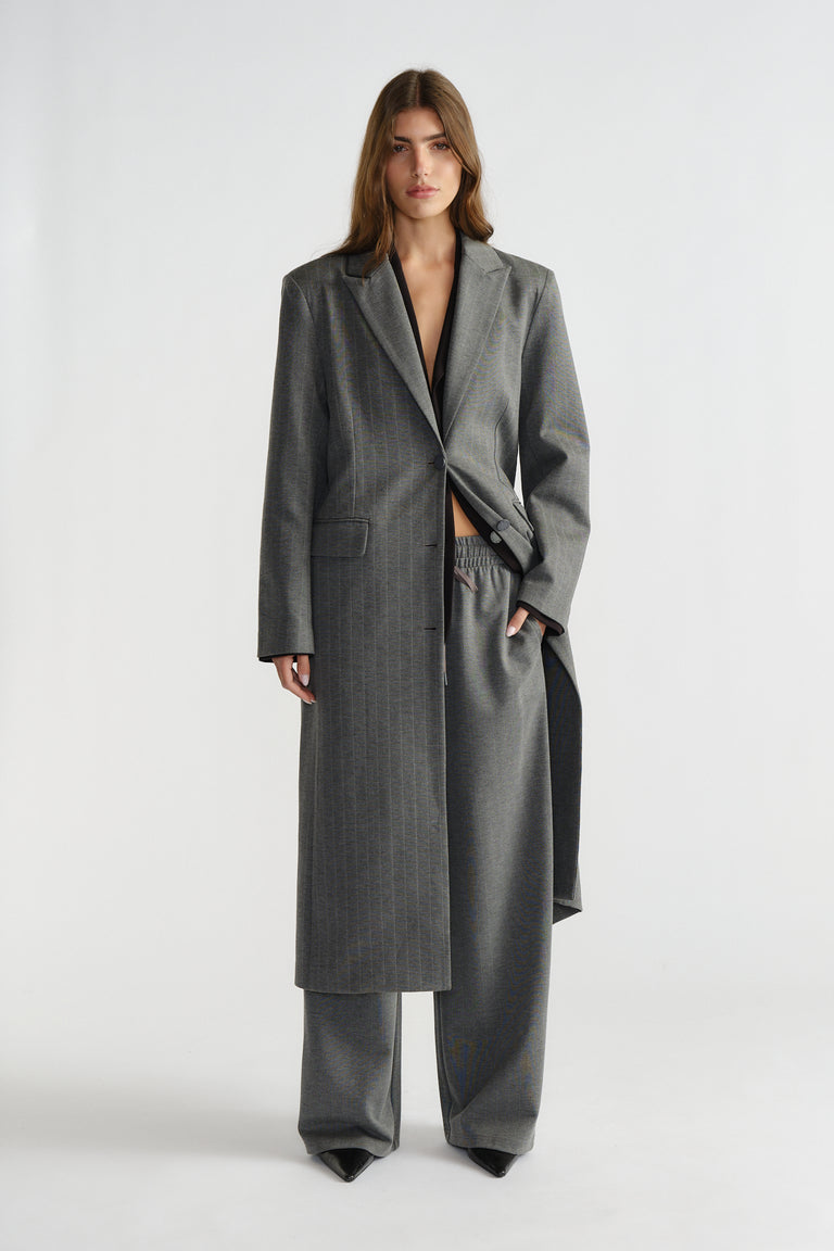 Woman wearing a charcoal grey pinstripe longline blazer over matching wide-leg pants in a minimalist studio setting.