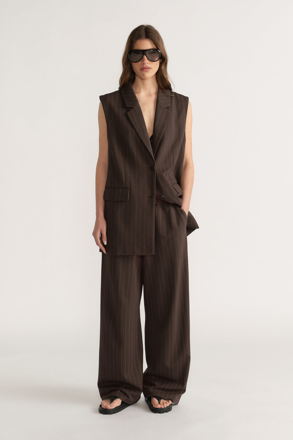 Woman wearing sleeveless brown pinstripe blazer and matching wide-leg pants with black sandals and sunglasses