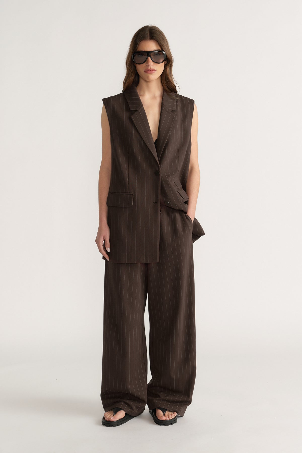 Woman wearing sleeveless brown pinstripe blazer and matching wide-leg pants with black sandals and sunglasses